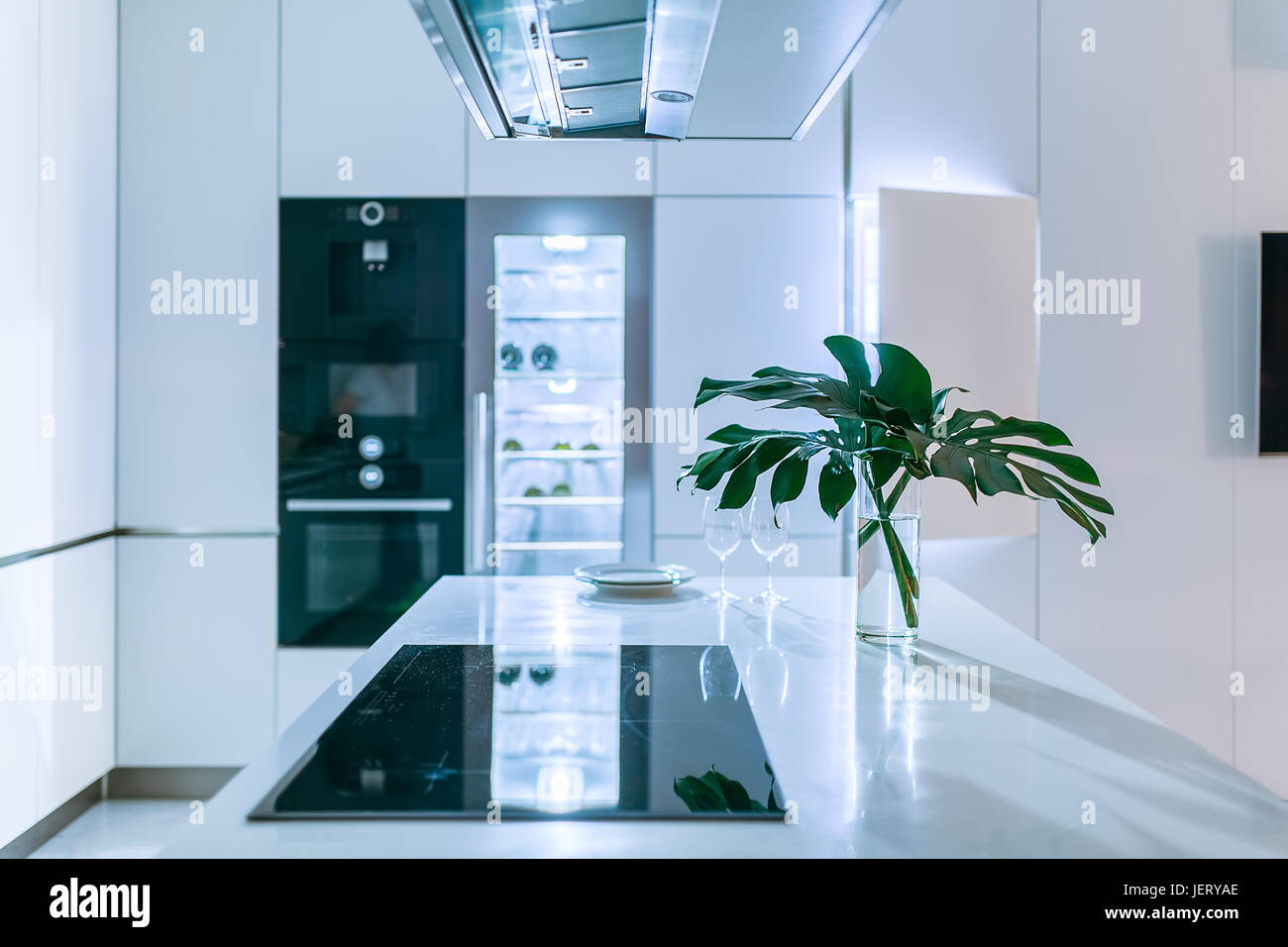 Illuminated modern kitchen with light walls. There is a white tabletop with a stove, plates, glasses, vase with green leaves. Behind it there is an ov Stock Photo