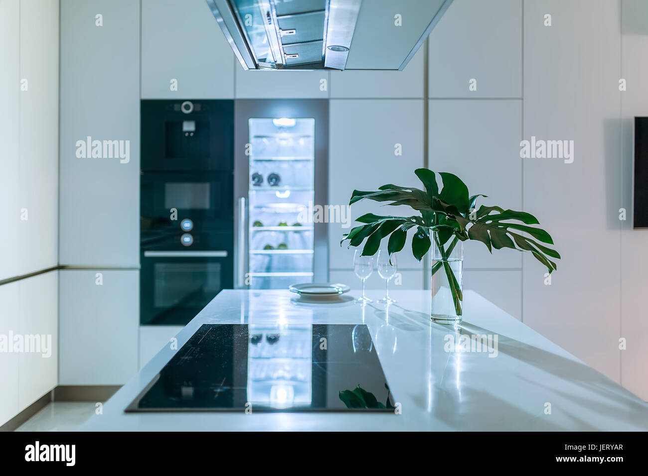 Luminous modern kitchen with light walls. There is a white tabletop with a stove, plates, glasses, vase with green leaves. Behind it there is an oven  Stock Photo