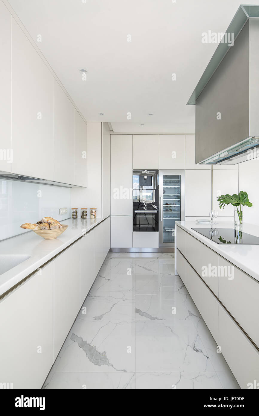 Modern white kitchen with light tiled floor. There are white lockers with a tabletop kitchen island with stove and kitchen hood leaves in a vase fr Stock Photo Alamy