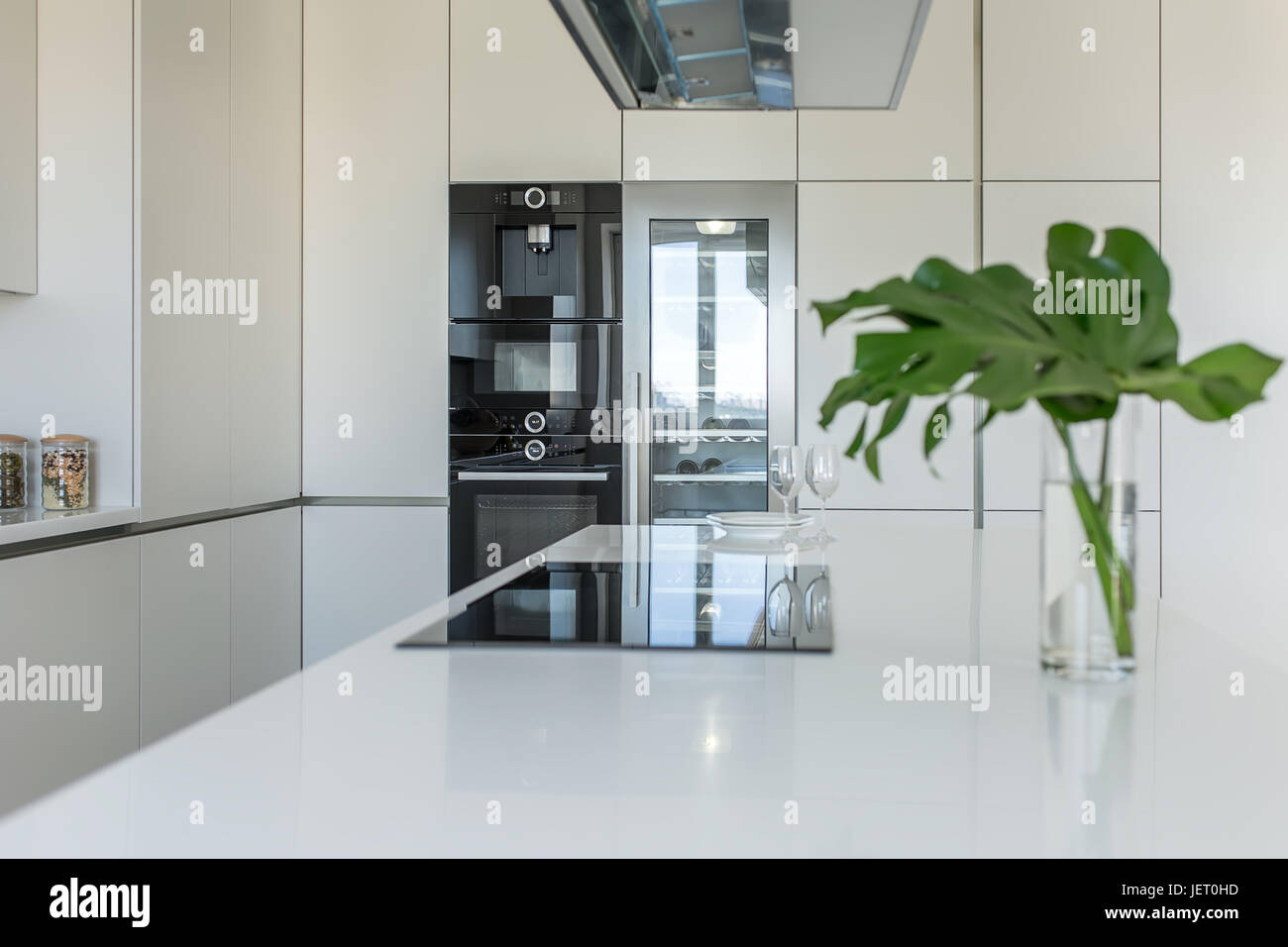Modern kitchen with light walls. There is a white tabletop with a stove, plates, glasses, vase with green leaves. Behind it there are lockers with an  Stock Photo