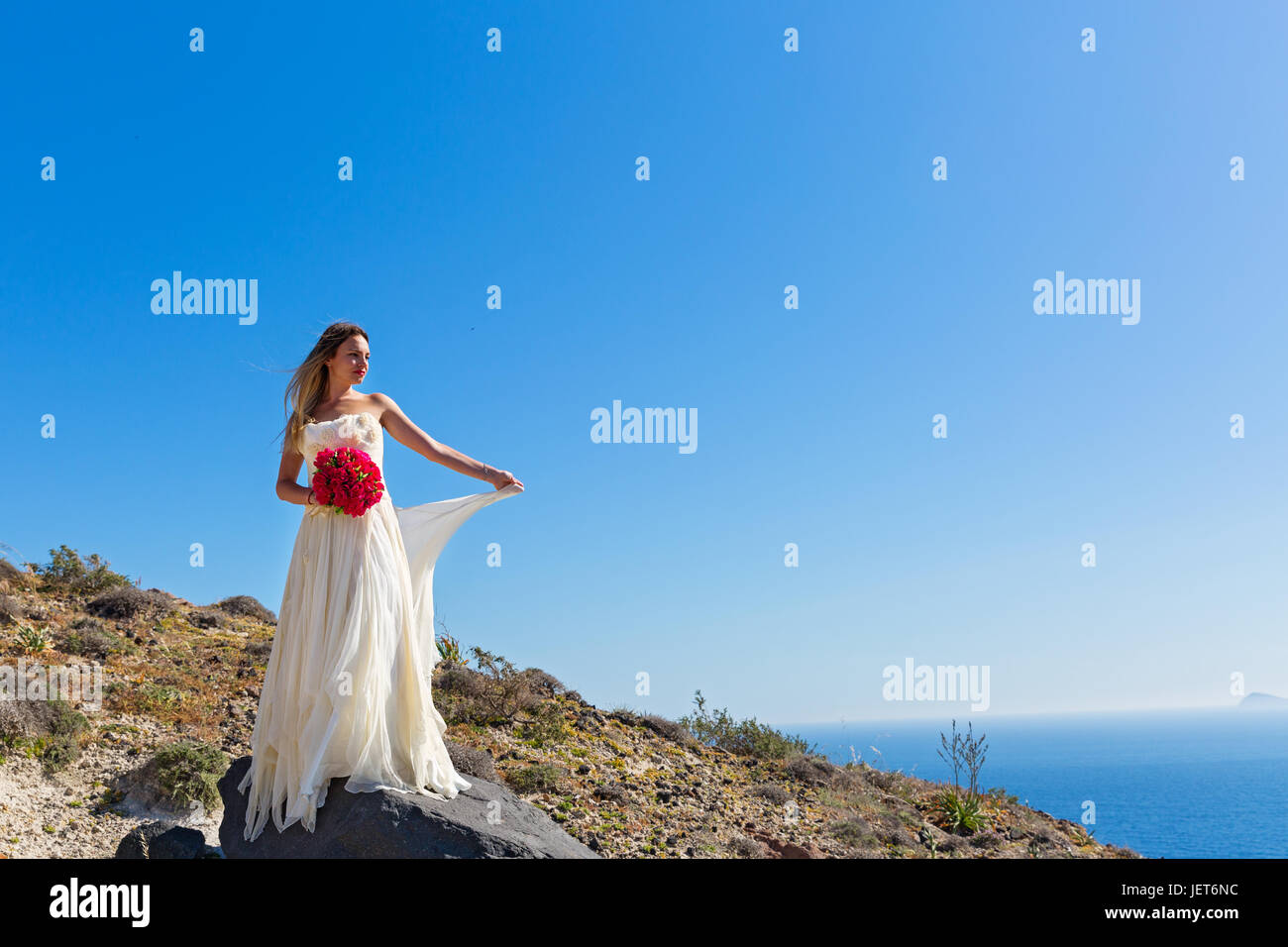 Beautiful woman in white dress stands on a high stone near the sea ...