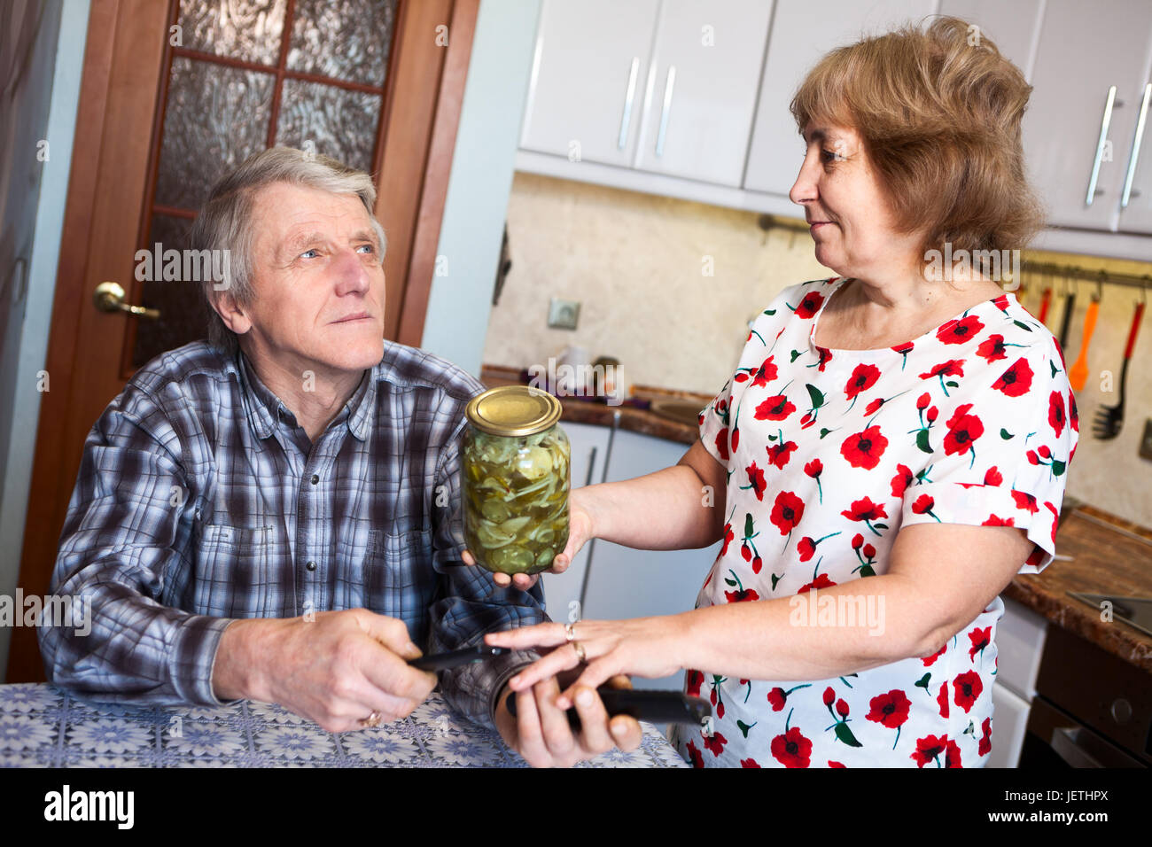 Mature woman asking busy husband to open the glass jar with marinated cucumbers while he watching tv Stock Photo