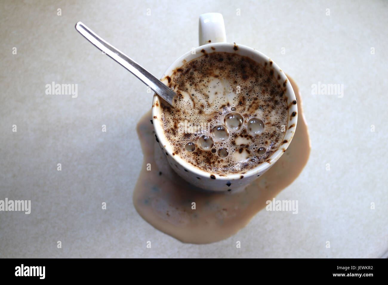 Mug or cup of coffee that has boiled over and spilt over onto the table or work top. Has the spoon in the mug. Coffee granule staining Stock Photo