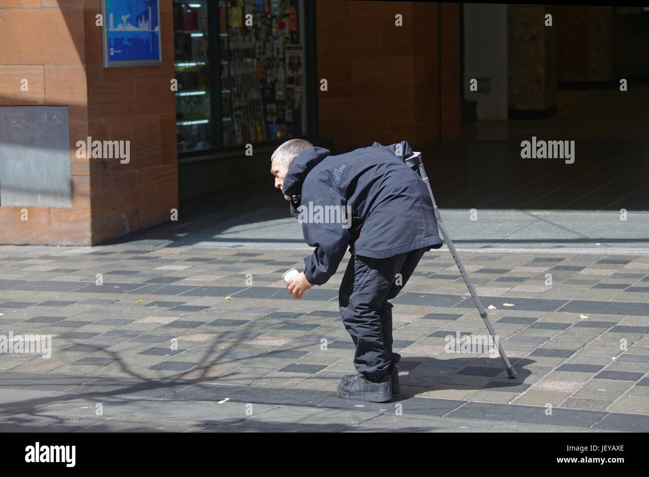 begging homeless unemployed man handicapped cripple with back disease and walking stick crooked with cup asking for money on street Stock Photo