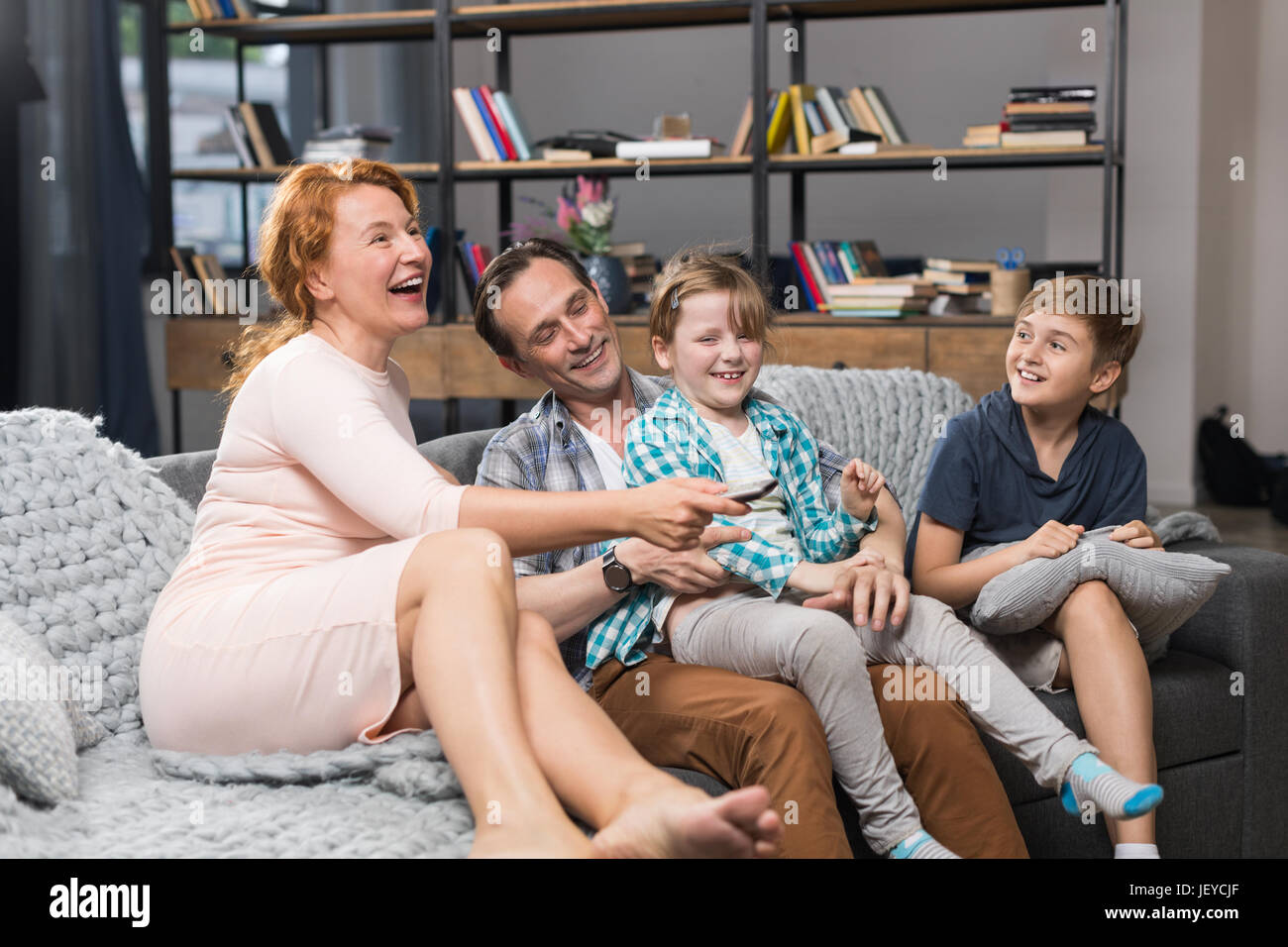 Family Sitting On Couch Watching TV, Happy Smiling Parents Spending Time With Children In Living Room Stock Photo