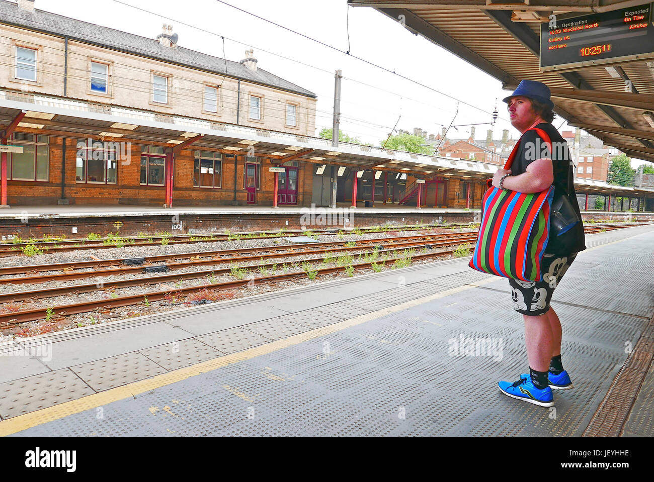 Young man stood alone on Preston railway station wearing blue trilby,blue trainers,Micky Mouse shorts and carrying a colourful striped bag Stock Photo