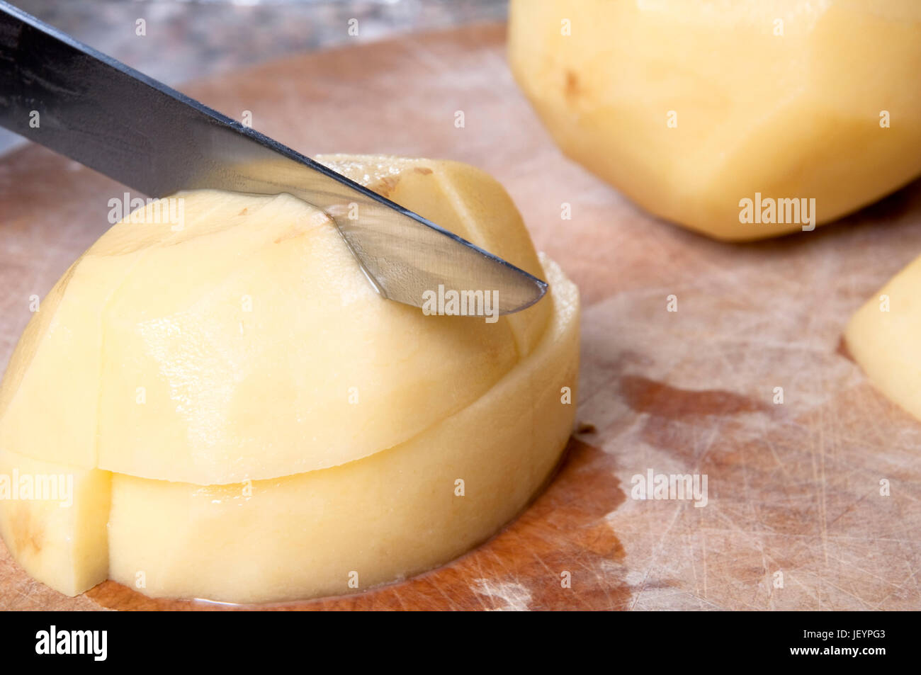 Close up action shot of potatoes being chopped on an old wooden chopping board, with visible cut marks.  Counter top visible in background. Stock Photo