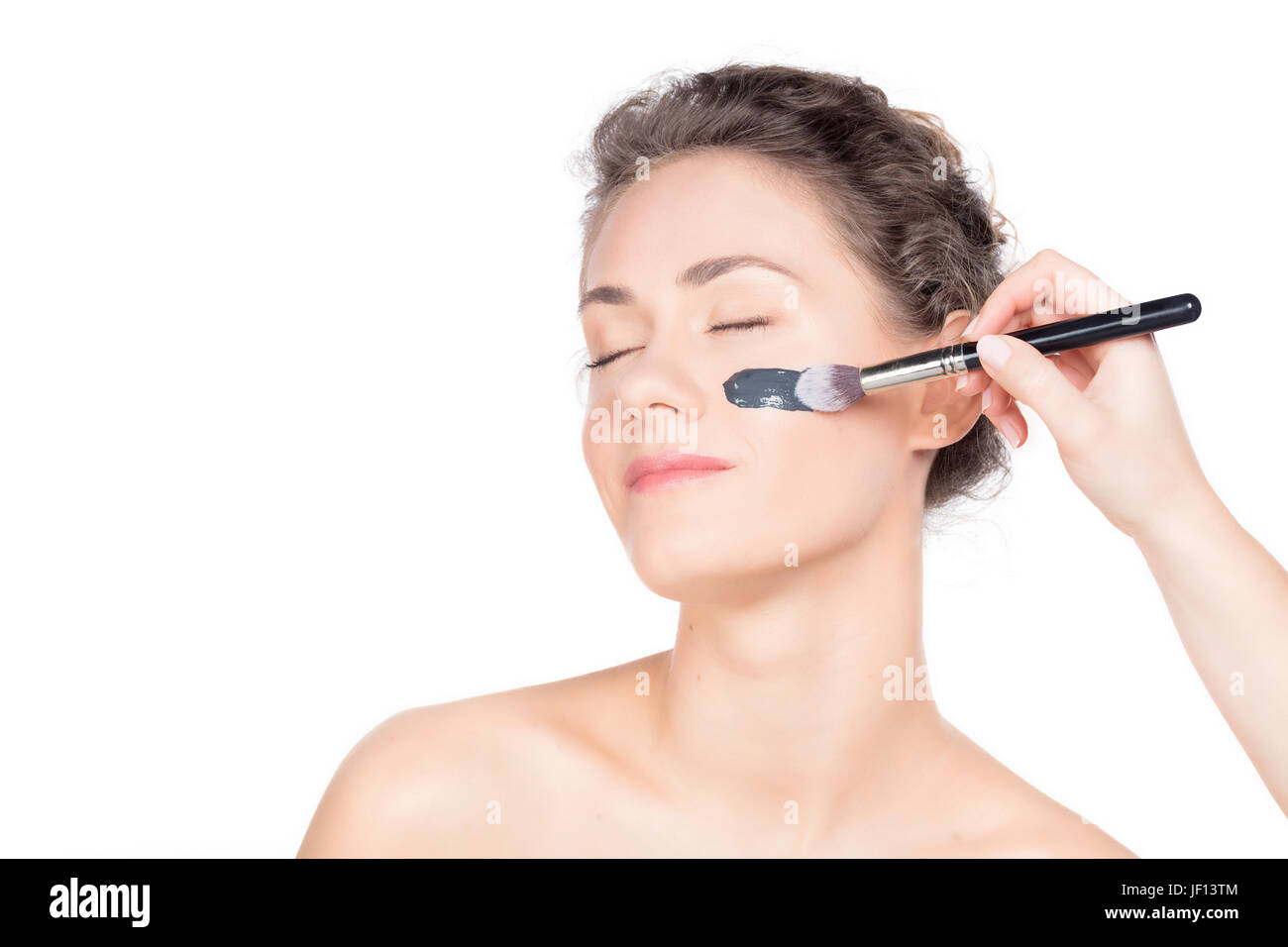 Young beautiful woman is having a black charcoal mask applied on her skin. Portrait close up on white background. Skincare and spa treatment. Stock Photo