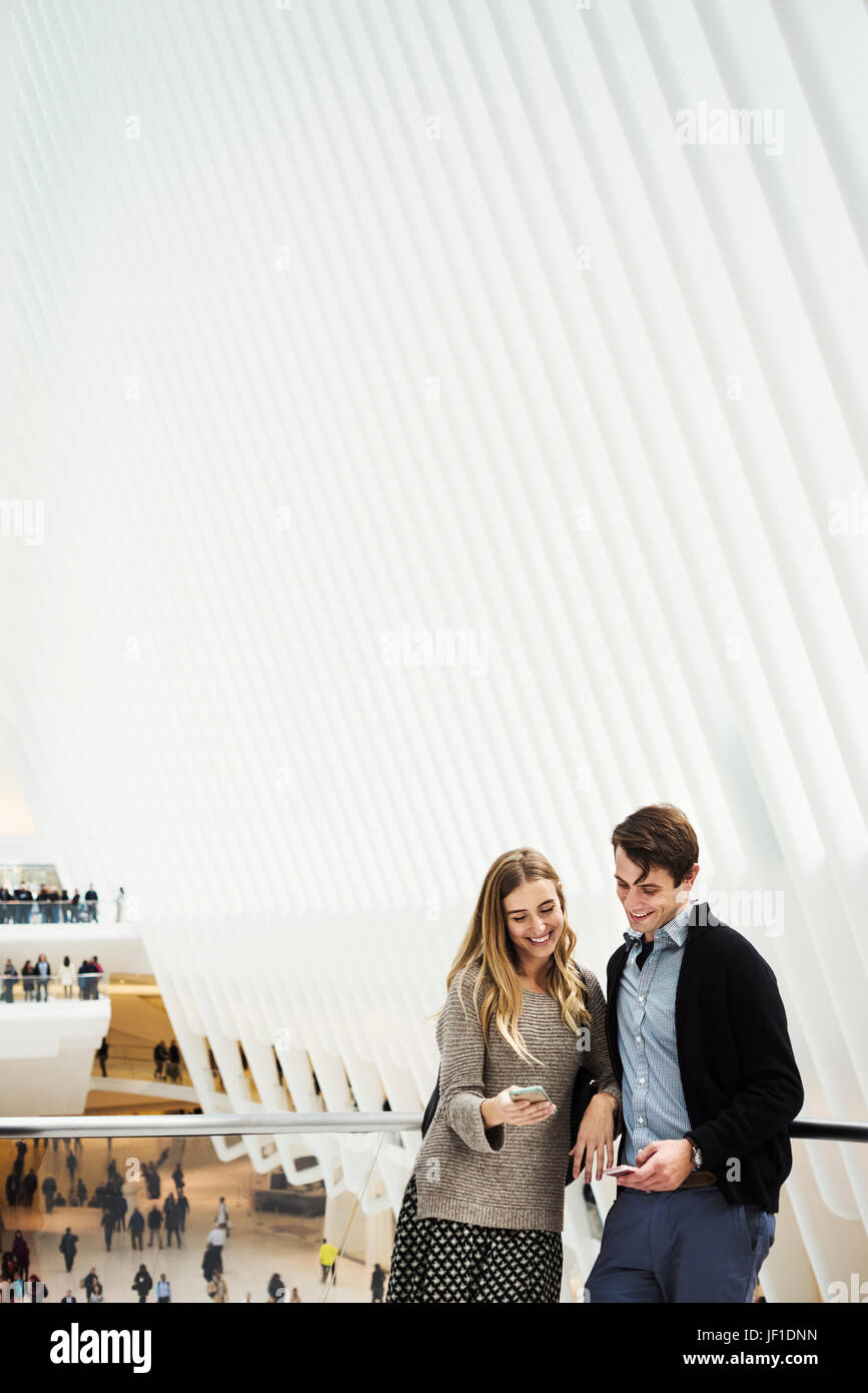 Two people standing on a balcony sharing a phone screen, with an overview of the central atrium of the World Trade Centre Oculus building and people p Stock Photo