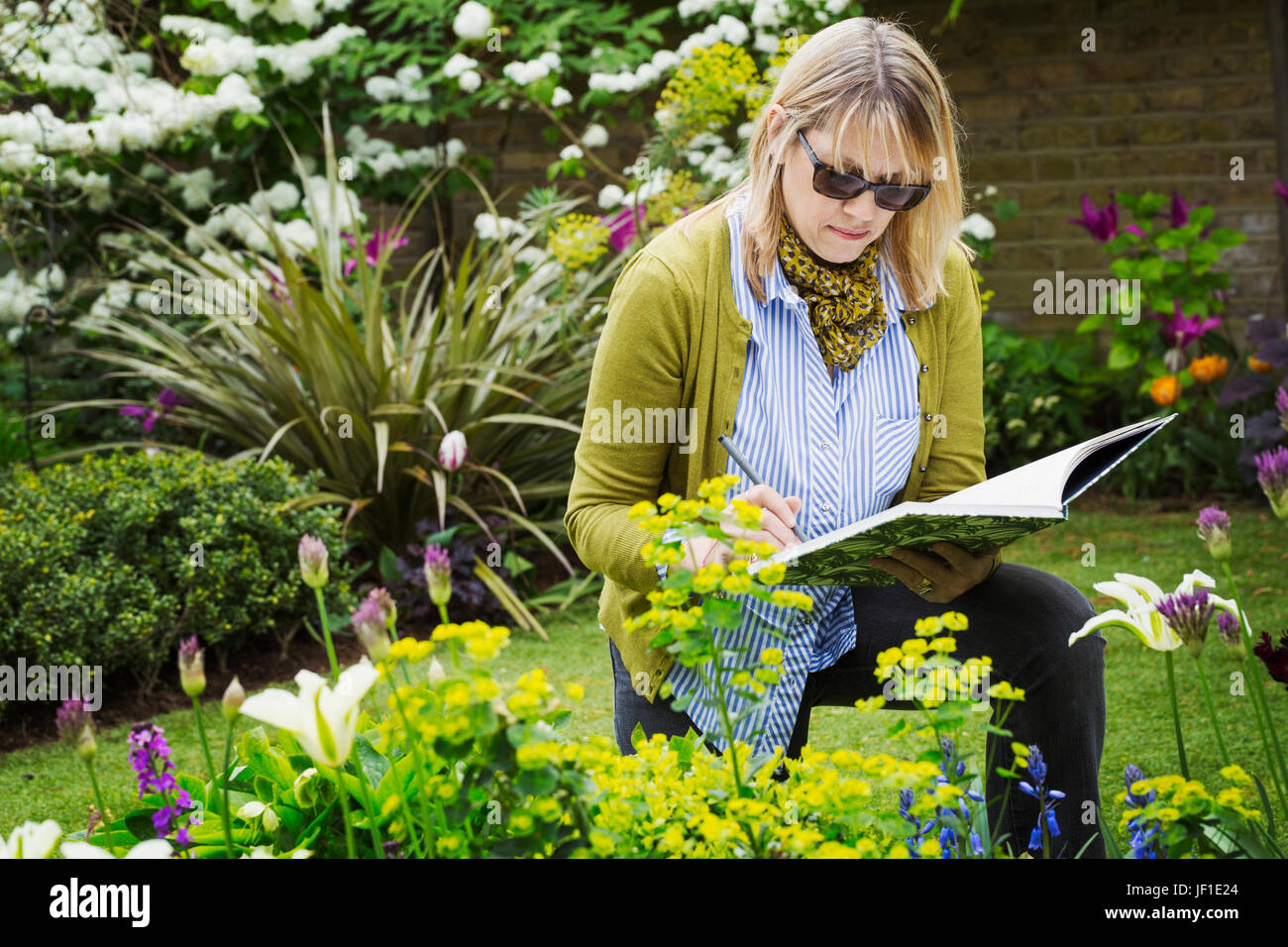 Woman wearing sunglasses standing in a garden by a flowerbed, drawing in a sketchbook. Stock Photo
