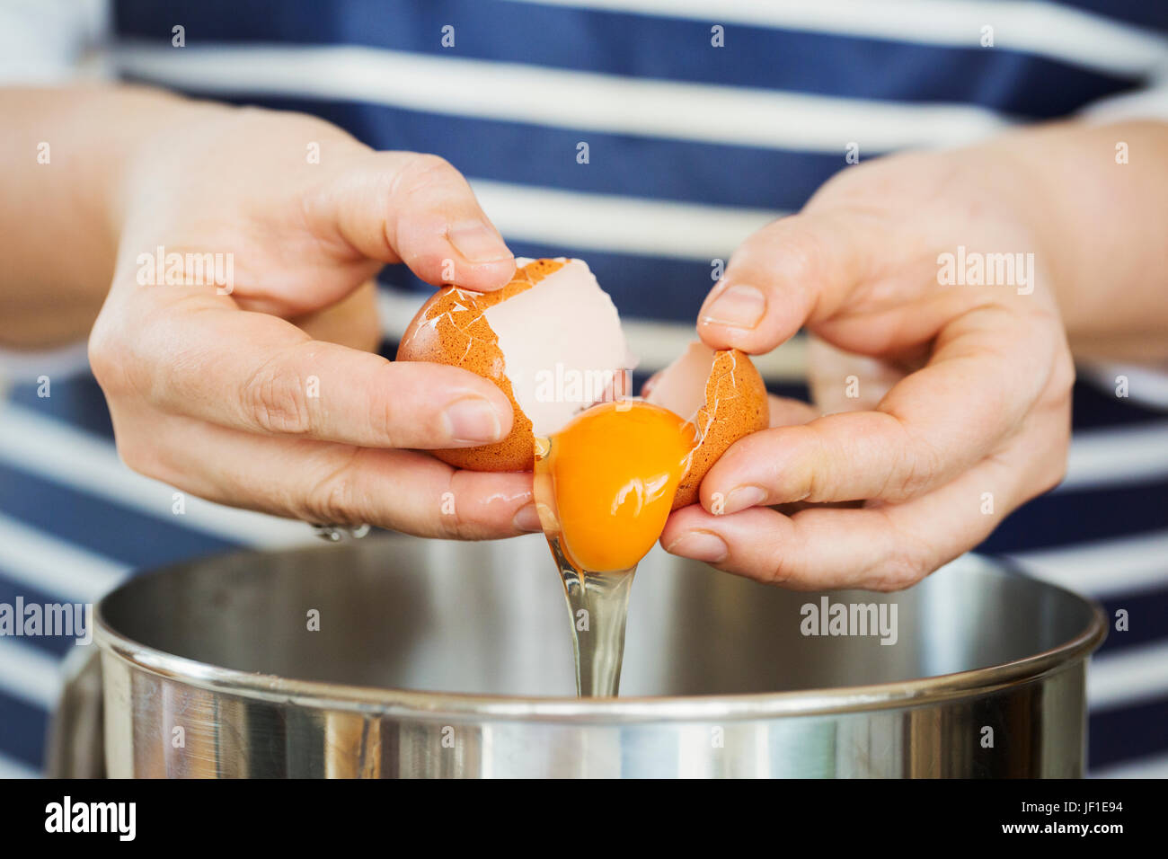 Close up of person wearing a blue and white stripy apron separating egg over a metal bowl. Stock Photo