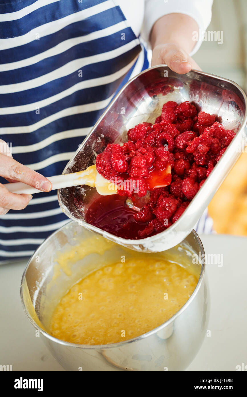 Close up high angle view of person wearing a blue and white stripy apron holding a metal dish, adding fresh raspberries to cake batter in a metal mixi Stock Photo