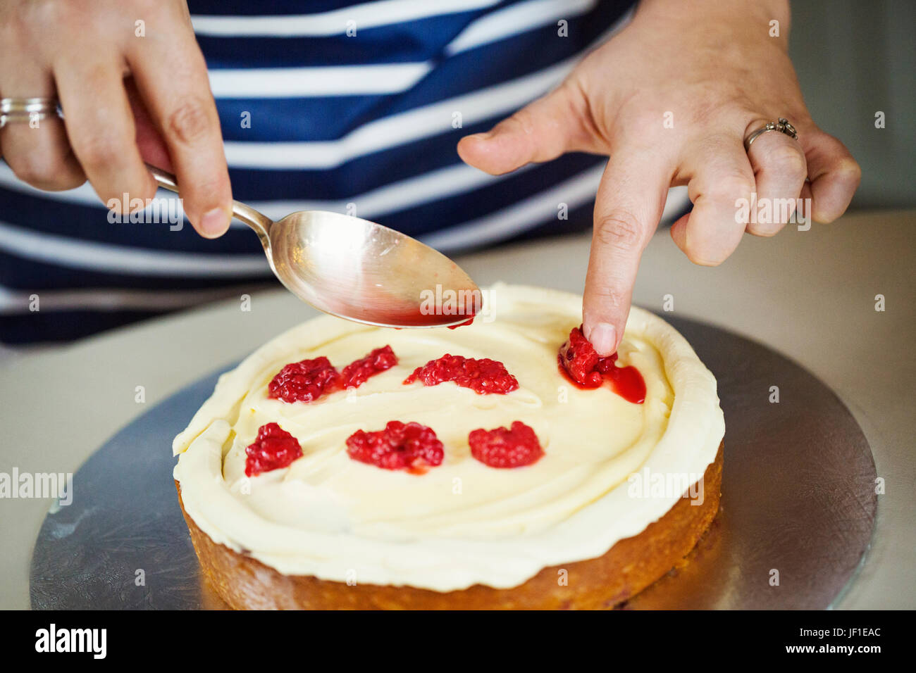 Close up high angle view of person wearing a blue and white stripy apron assembling a layer cake, holding spoon, placing raspberries on layer of cream Stock Photo