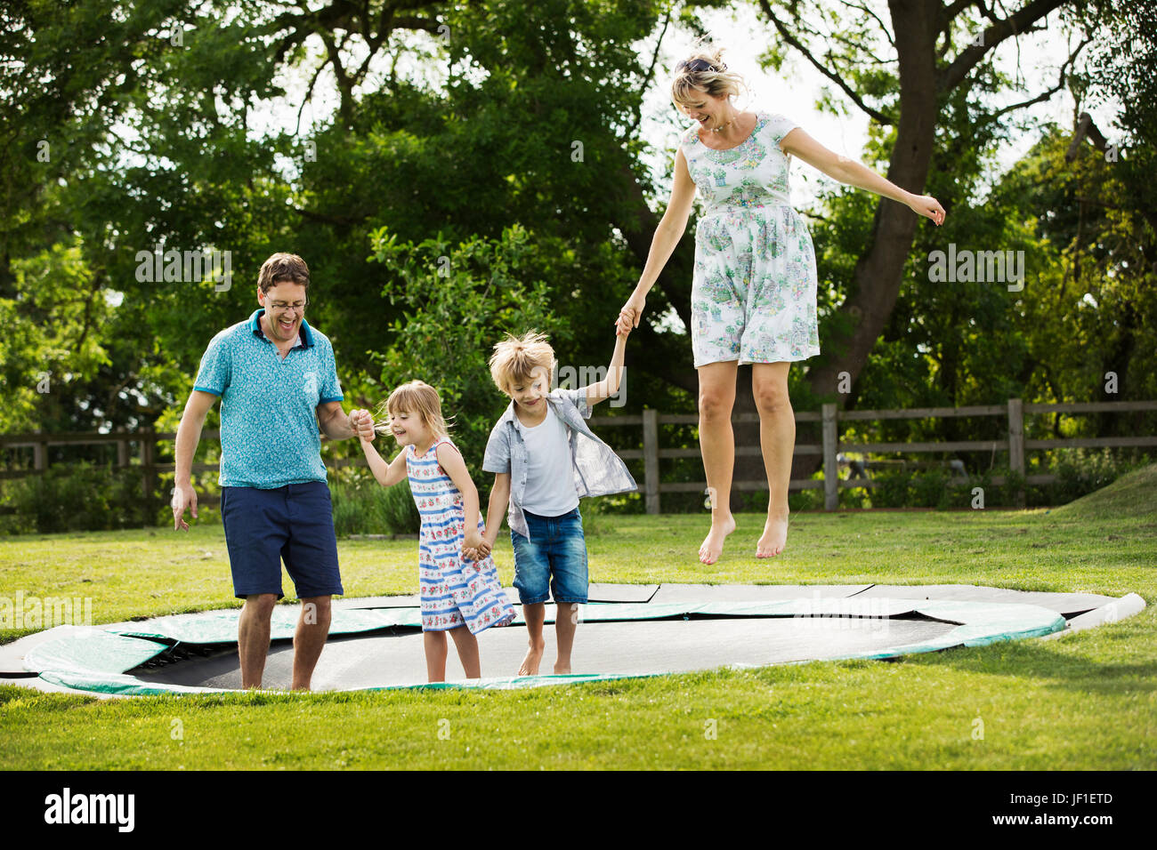 Man, woman, boy and girl holding hands, jumping on a trampoline set in the lawn in a garden. Stock Photo
