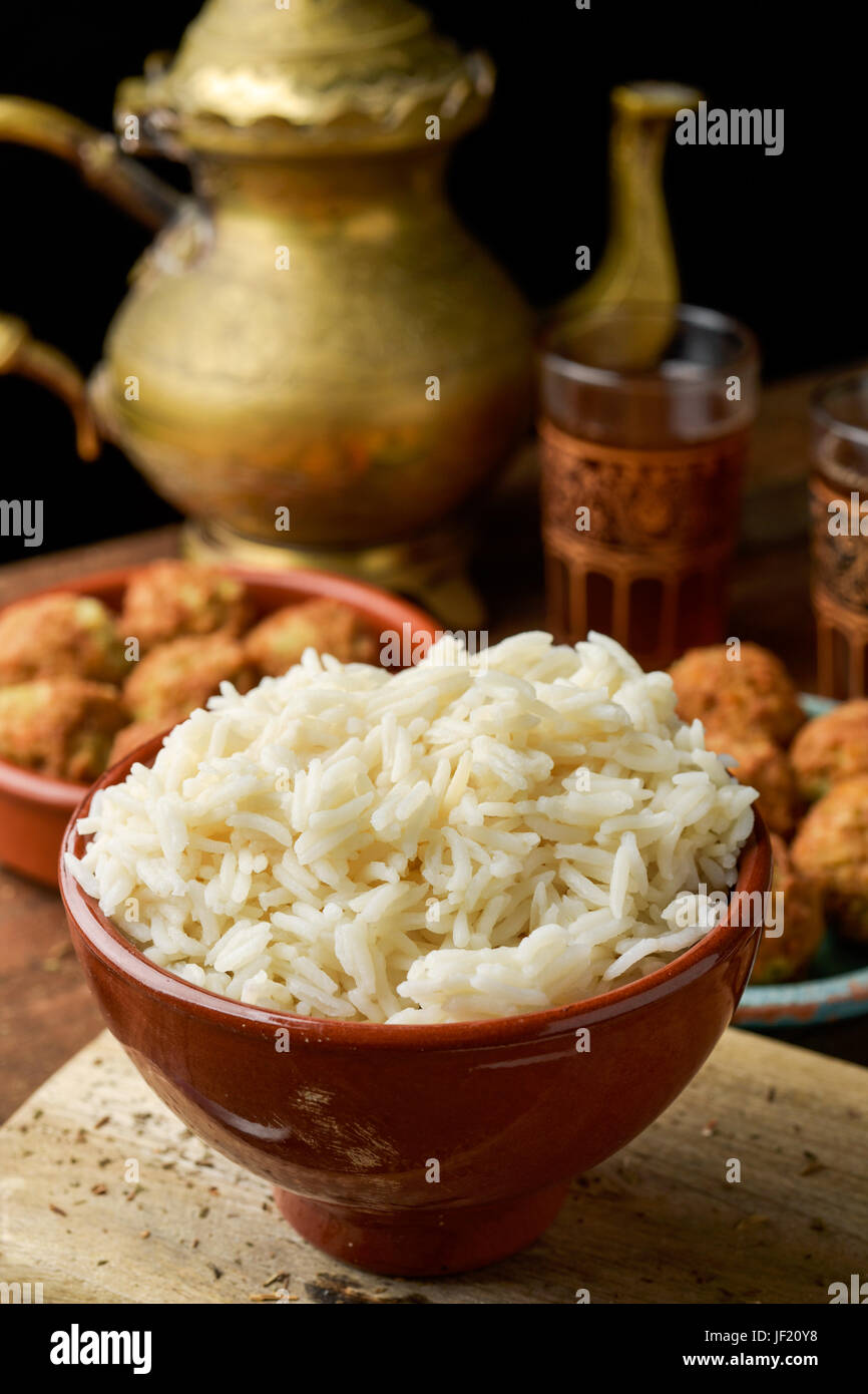 closeup of an earthenware bowl with steamed long grain rice and some plates with falafel on a rustic wooden table, with a golden teapot and some ornam Stock Photo