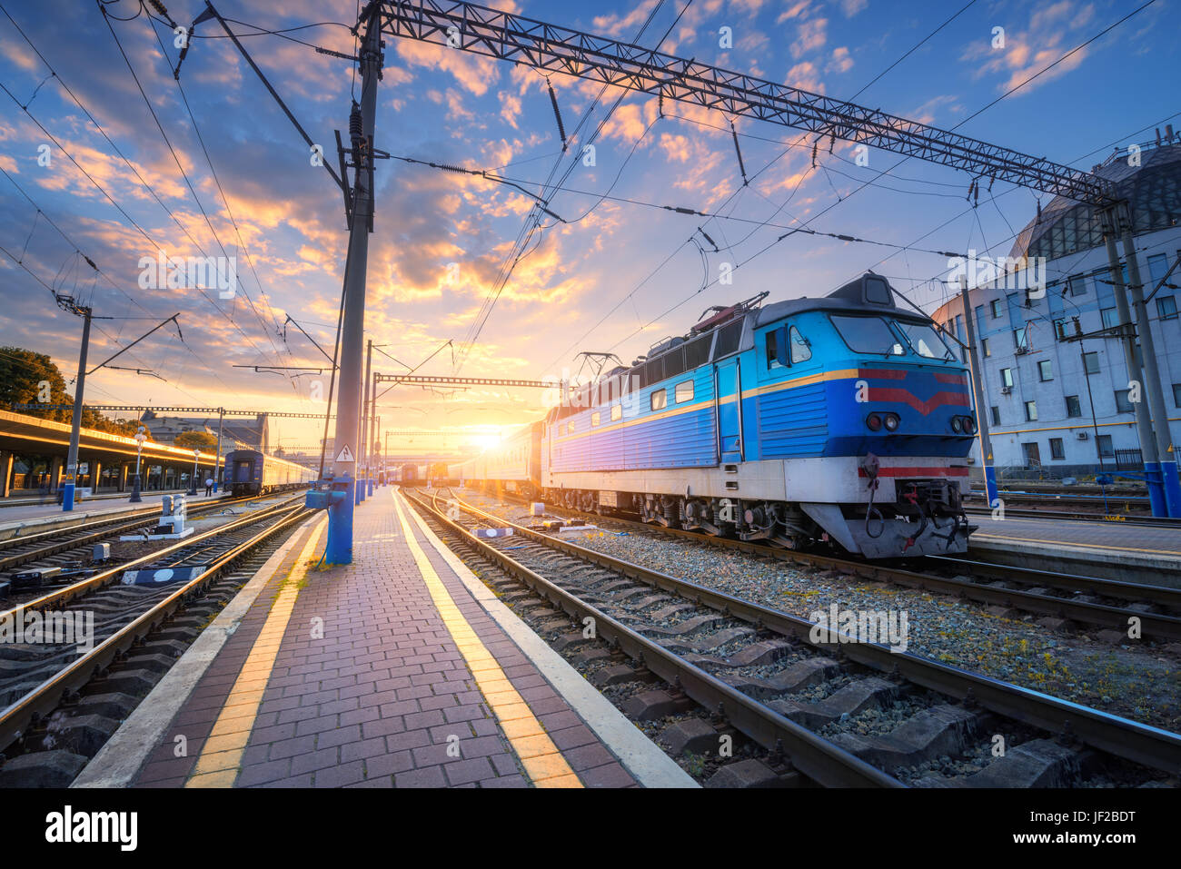 Blue train at the railway station at sunset. Amazing industrial landscape with old locomotive, old buildings, rails and colorful sunset sky with cloud Stock Photo