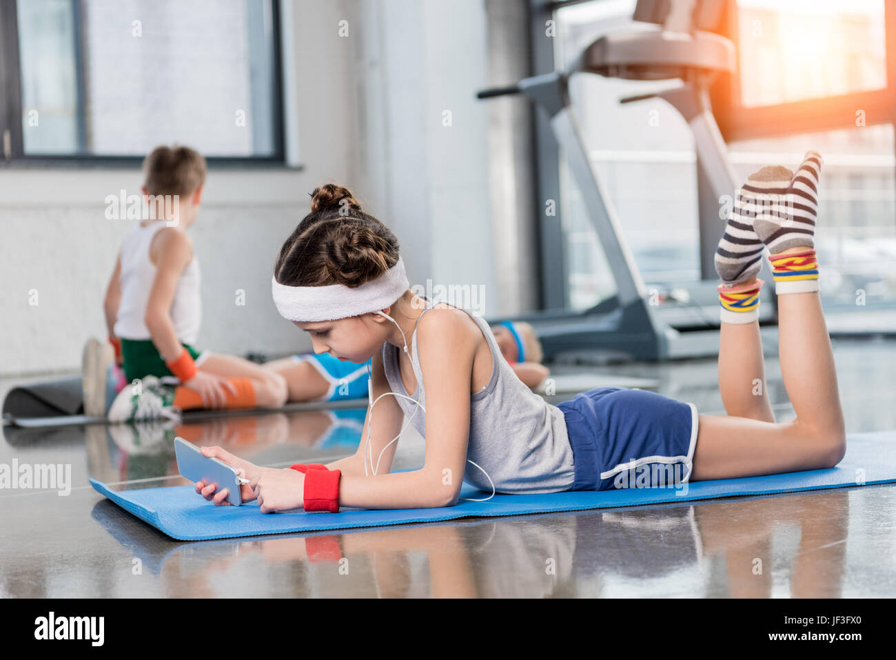 Side view of little girl in earphones lying on yoga mat and using smartphone in gym, children sport school concept Stock Photo