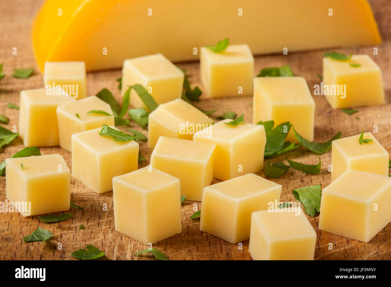 Close up of cubes of yellow cheese with herbs stacked randomly on wooden chopping board Stock Photo