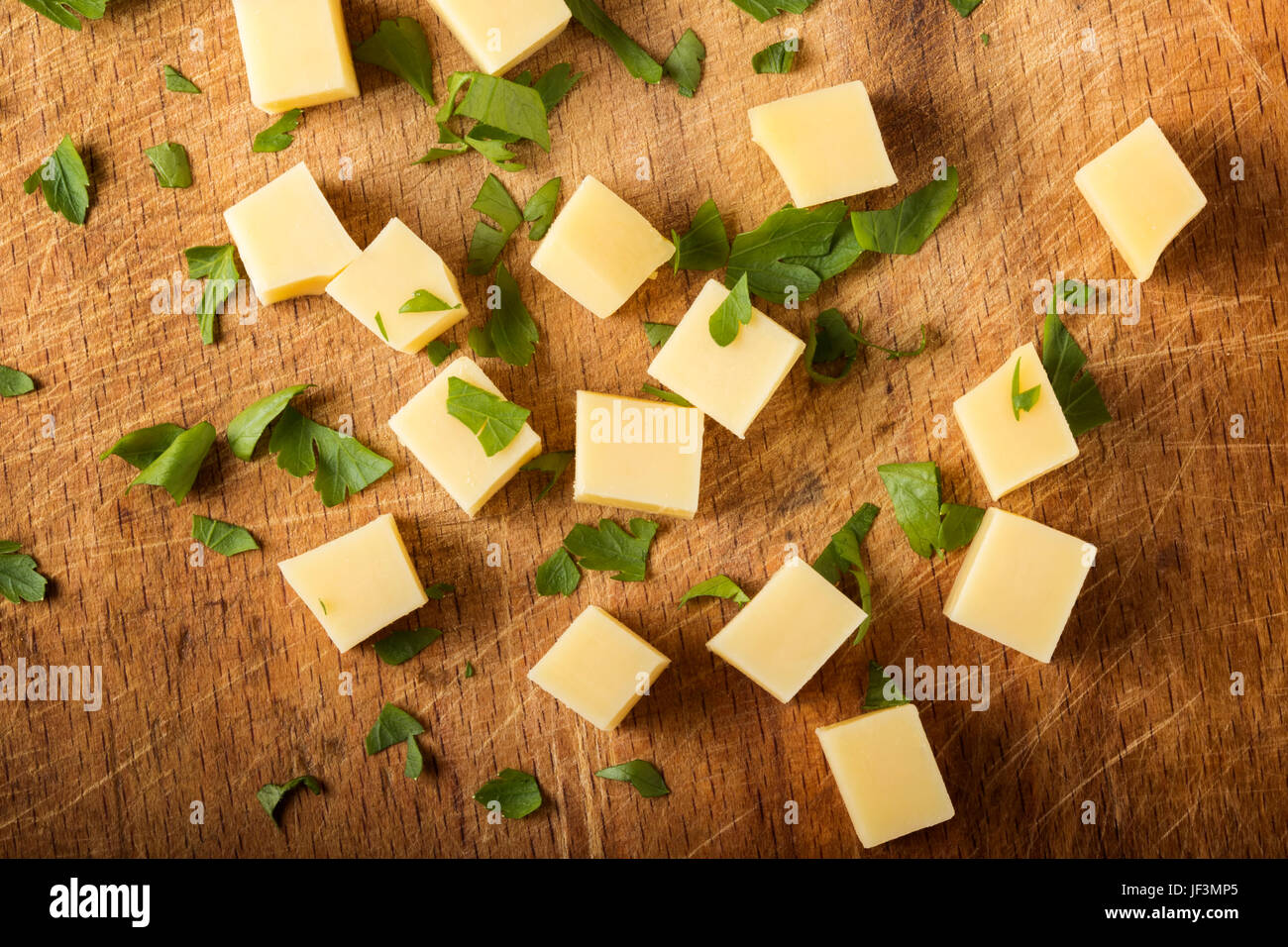 Cubes of yellow cheese with herbs stacked randomly on wooden chopping board Stock Photo