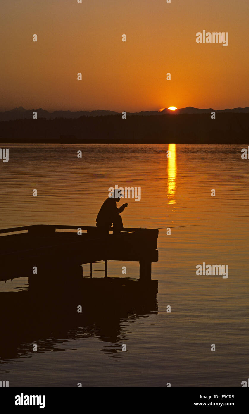 Sunrise with older man silhouetted fishing off dock on lake Washington with Cascade Mountains Seattle Washington State USA Stock Photo