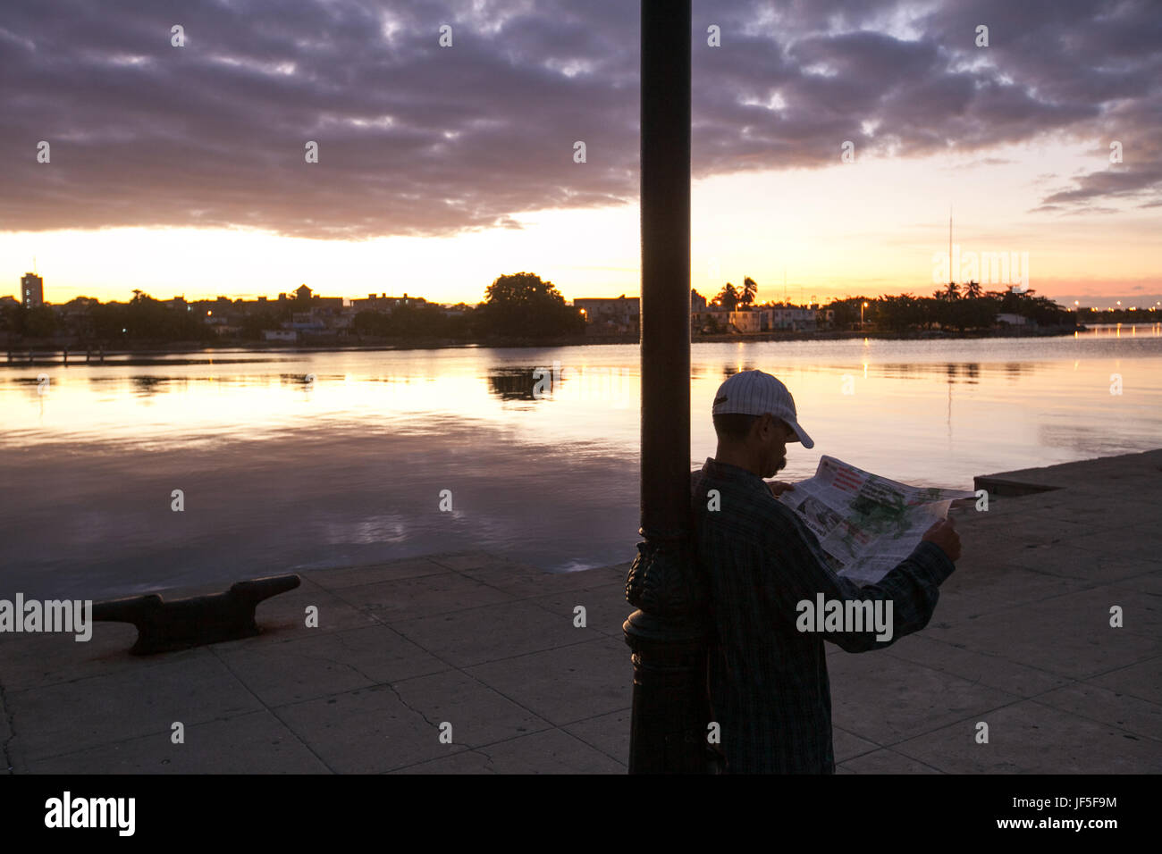 Along the waterfront of Cienfuegos, a man leans again a lamp post and reads a newspaper at dusk. Stock Photo