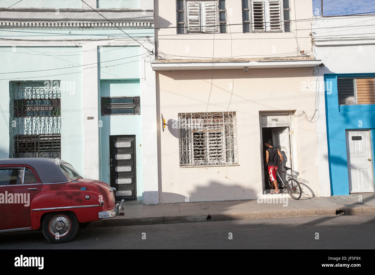 Near a classic American car, a young man walks his bicycle into his home on the streets of Cienfuegos. Stock Photo