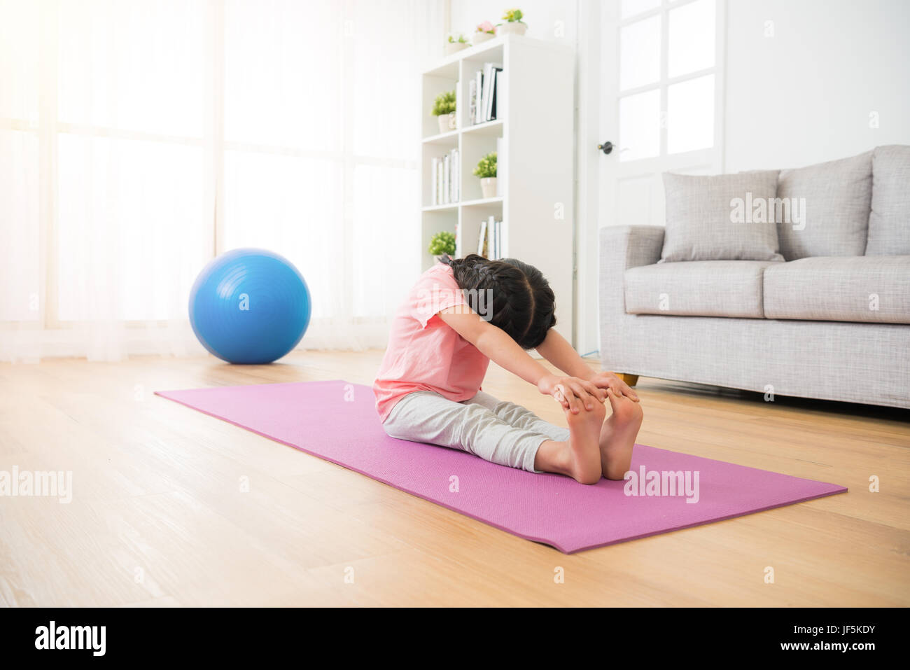asian little youth girl sitting on the wooden floor with mat straighten her legs and hands catch feet stretch body  with yoga fit sport at home in the Stock Photo
