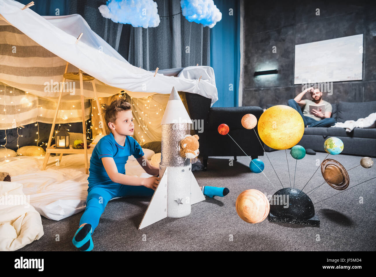 boy sitting in blanket fort with toy rocket and solar system model, father sitting on sofa behind Stock Photo