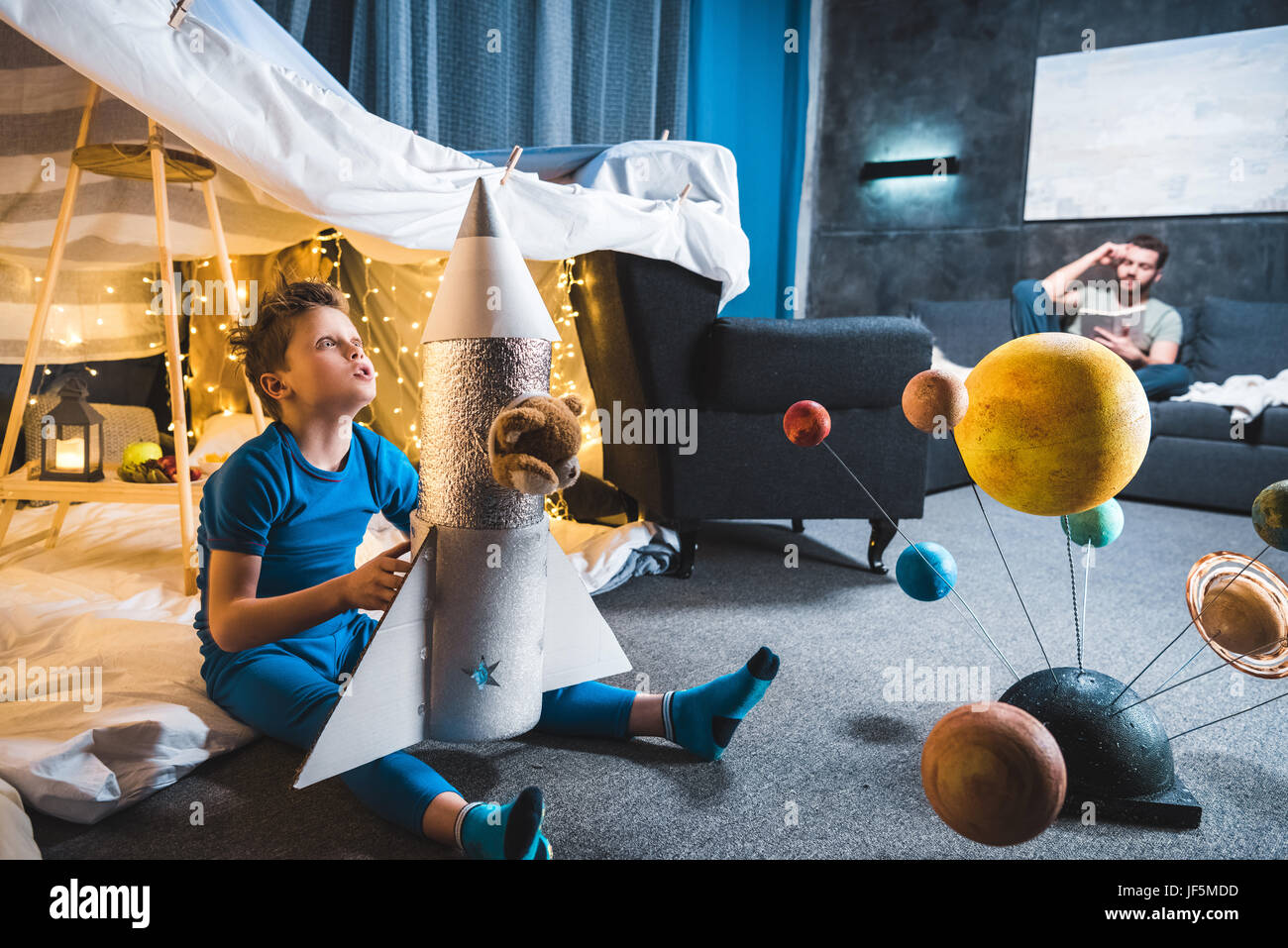 boy sitting in blanket fort with toy rocket and solar system model, father sitting on sofa behind Stock Photo