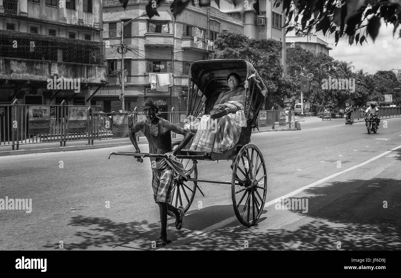 Man pulls woman passenger in wooden rickshaw along tarmac street flanked by tenement blocks in Kolkata, India. Stock Photo