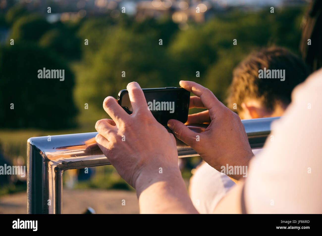 Man holding a smartphone in hands, leaning on metal railing, stabilising to take a picture on the phone. Stock Photo