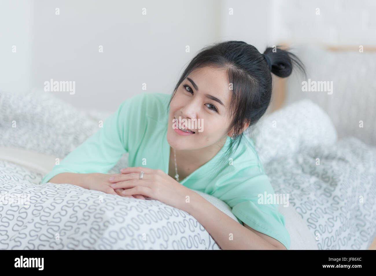 Young attractive Asian woman wear green casual cloth smiling while laying down on bed. Lazy girl activity on sunday morning. Stock Photo