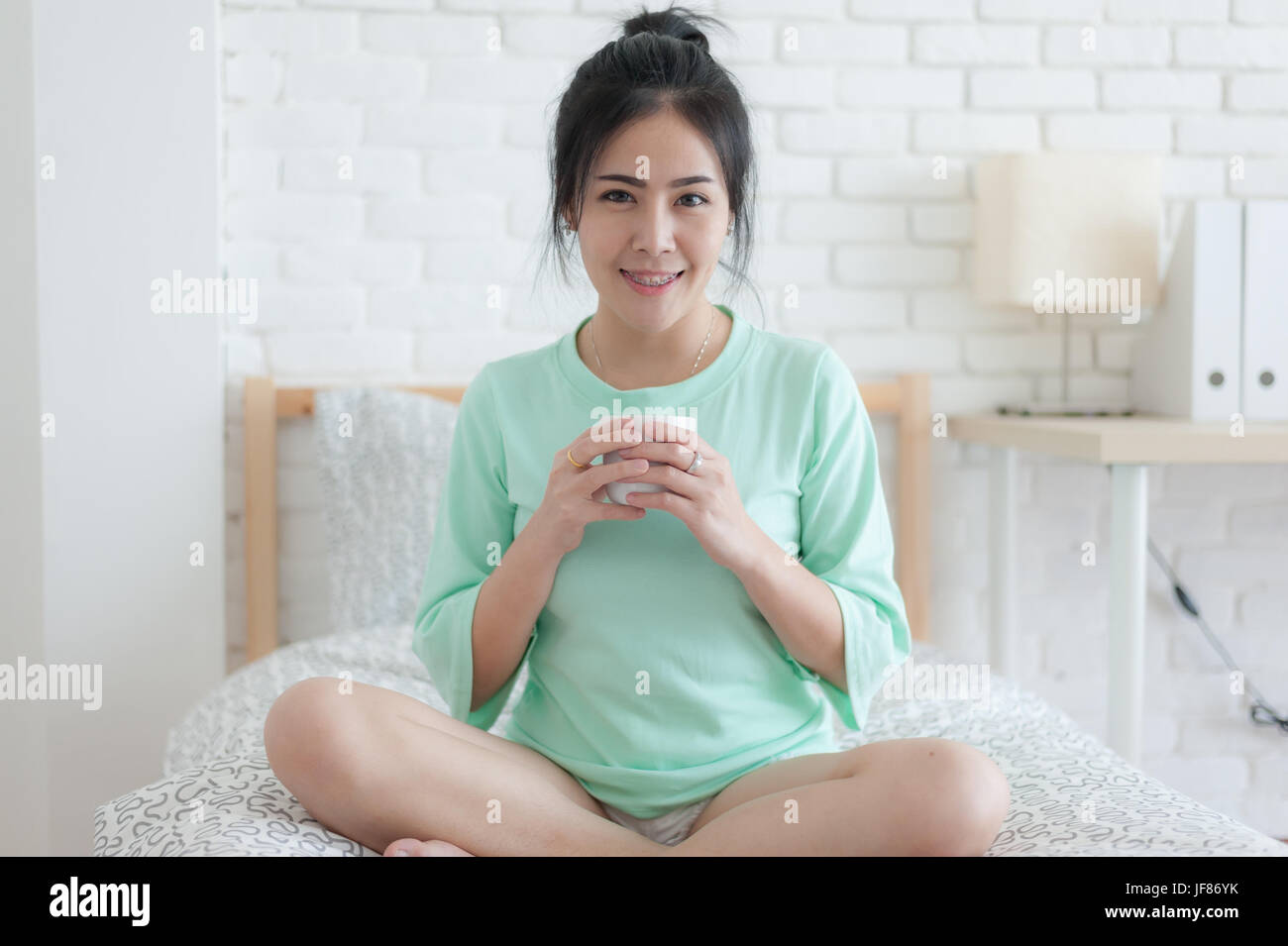 Young attractive Asian woman wear green casual cloth holding coffee cup while sitting on bed. Lazy girl activity with coffee on sunday morning. Stock Photo