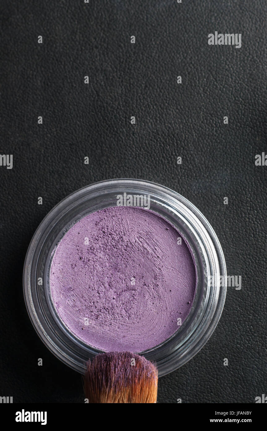 Overhead close up (macro) of an opened pot of purple eye shadow, with make up brush coated in powder on black leatherette background. Stock Photo