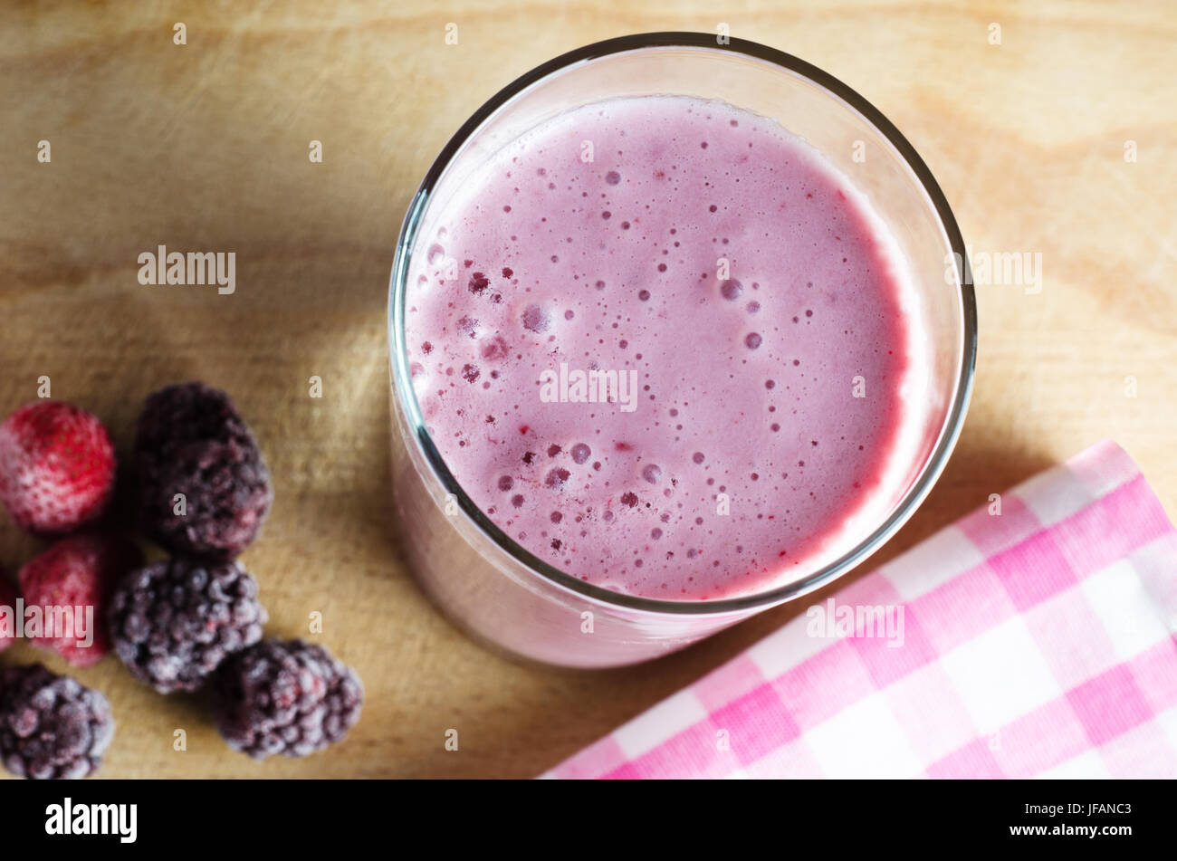 Overhead close up of a pink smoothie (made with soya milk)  on an old wooden chopping board with frozen Summer berries to the left and a gingham check Stock Photo