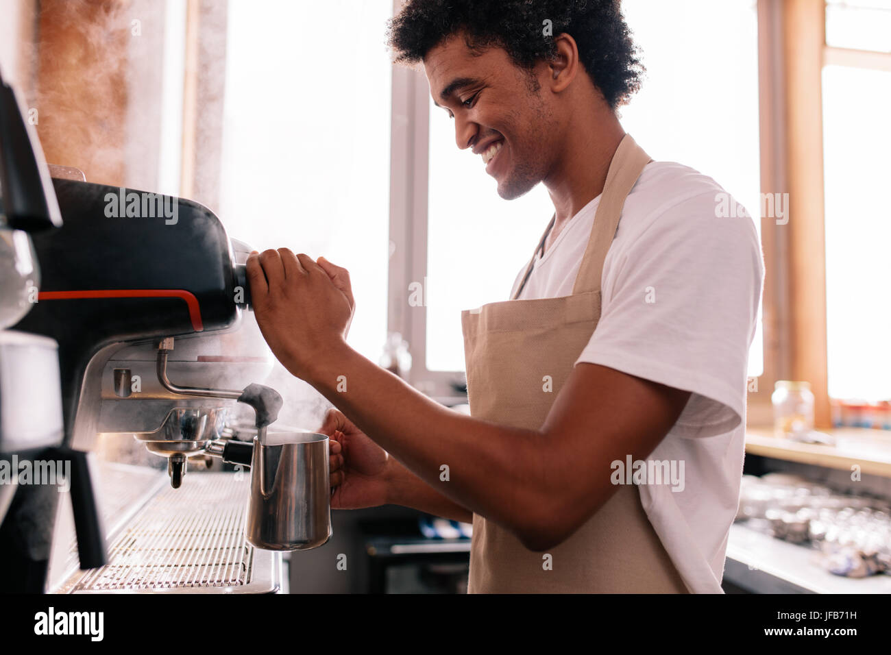 Smiling barista holding metal jug warming milk using the coffee machine. Happy young man preparing coffee at counter. Stock Photo