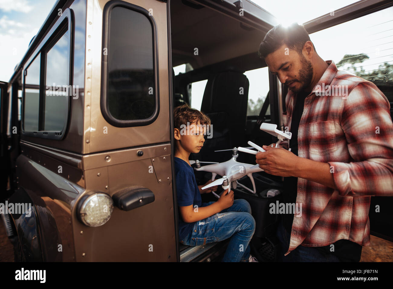 Shot of father and son at the back of their car setting up a drone to fly. Young man and son assembling a drone on road trip. Stock Photo