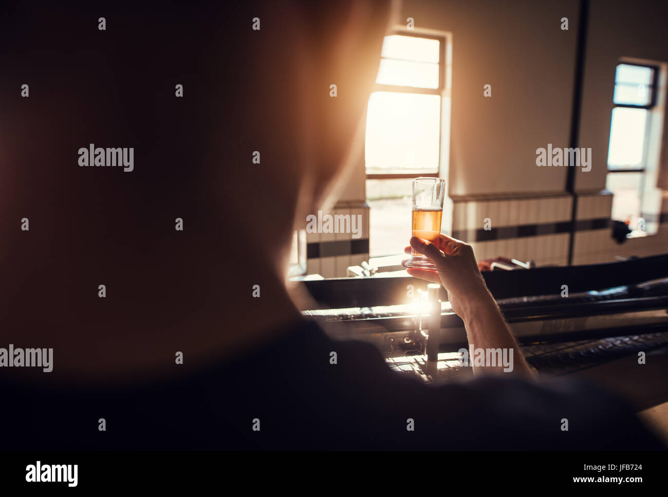 Rear view of manufacturer examining beer in glass at brewery. Man working at brewery checking the quality of beer in factory. Stock Photo