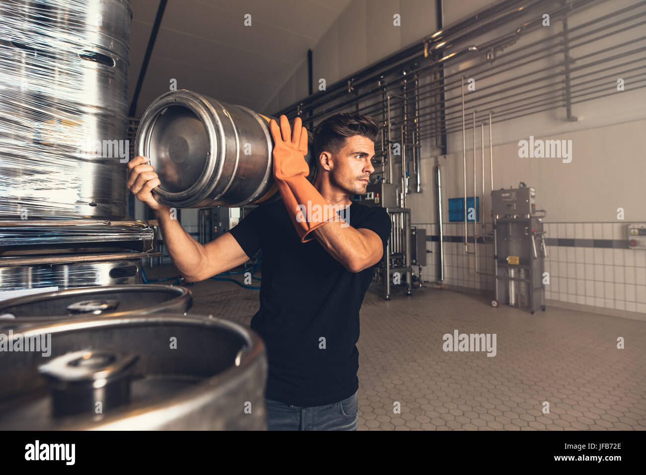 Brewer with large metal container at brewery factory. Young man working at warehouse in brewery. Arranging metal beer barrel at storage area. Stock Photo
