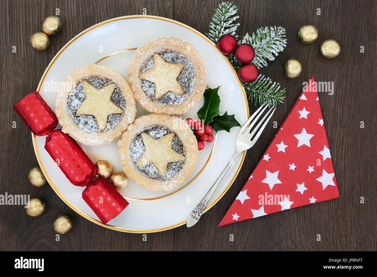 Christmas mince pie tarts on a plate with sliver fork, napkin and cracker with bauble decorations and foil wrapped chocolates on oak background. Stock Photo