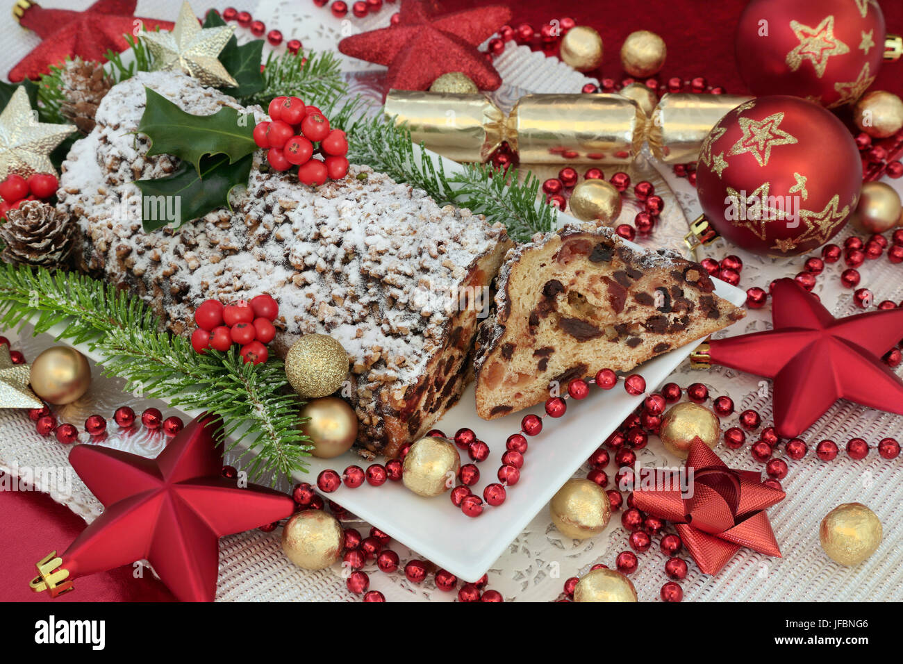 Traditional christmas chocolate stollen cake on a plate with slice, red and gold bauble decorations, foil wrapped chocolates, holy and fir. Stock Photo