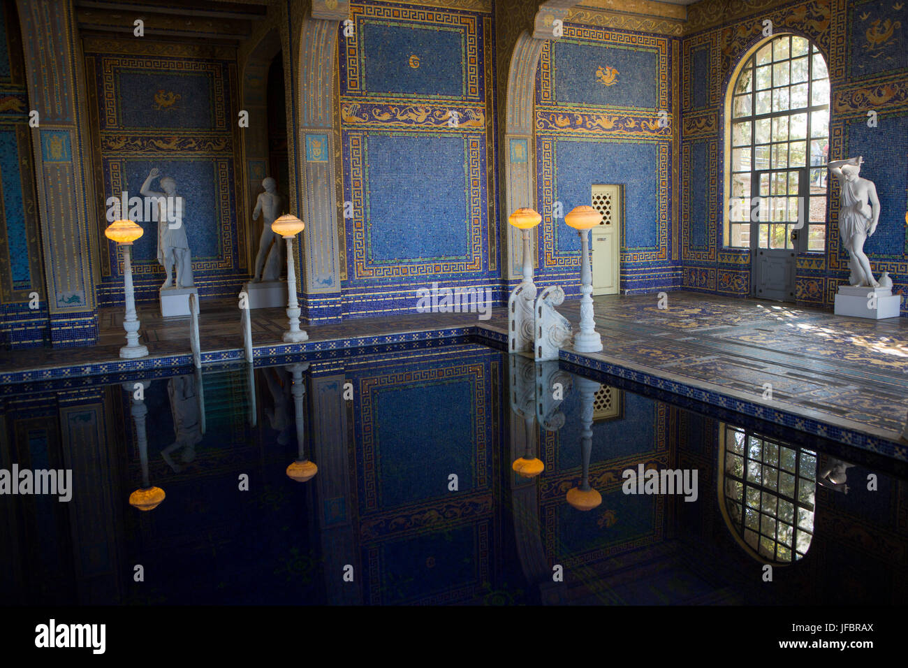 The Roman Pool at Hearst Castle, styled after an ancient Roman bath, is tiled with mosaic patterns in blue and gold, lit with ornate lighting, and surrounded by sculptures. Stock Photo