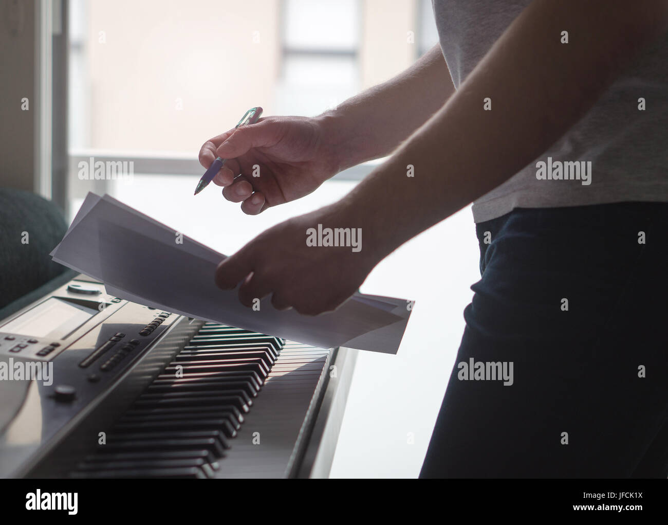 Musician brainstorming and innovating new song ideas at piano by the window. Composer writing notes to paper or planning arrangement. Stock Photo