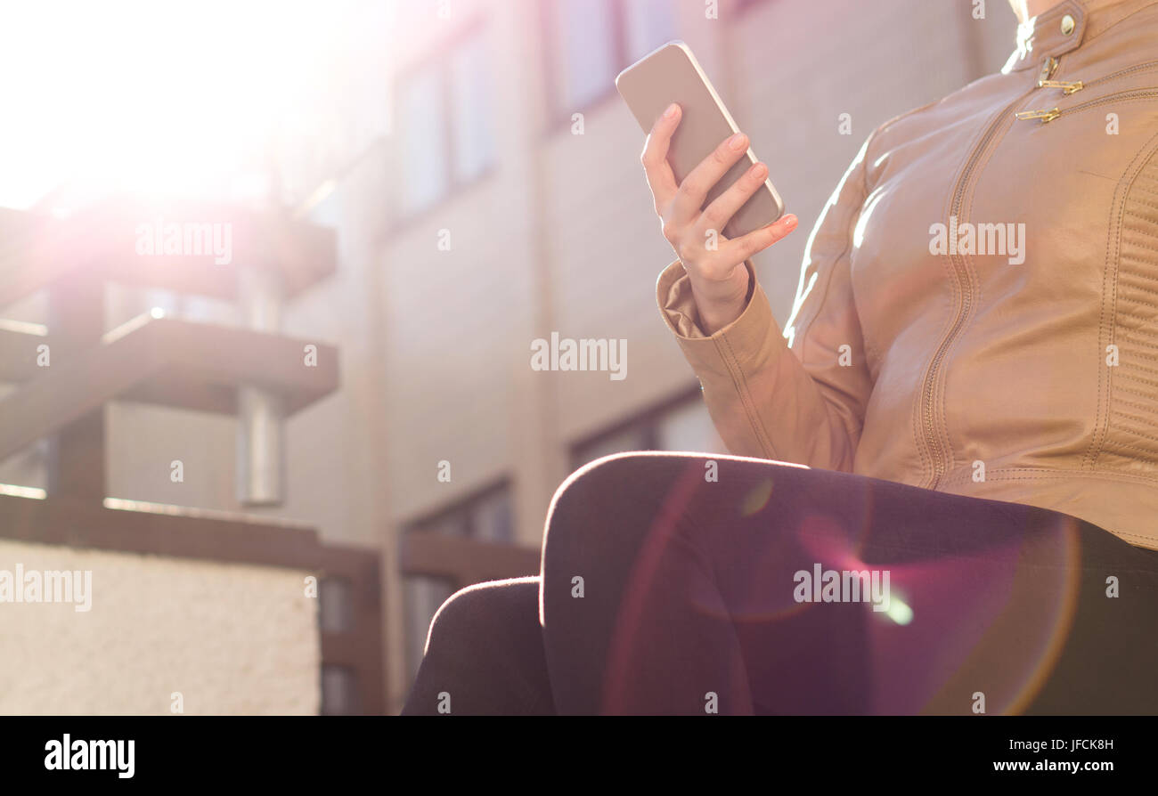 Relaxed girl using mobile phone in sunshine. Stylish young woman with trendy clothes touching smartphone and chilling. Modern urban lifestyle. Stock Photo