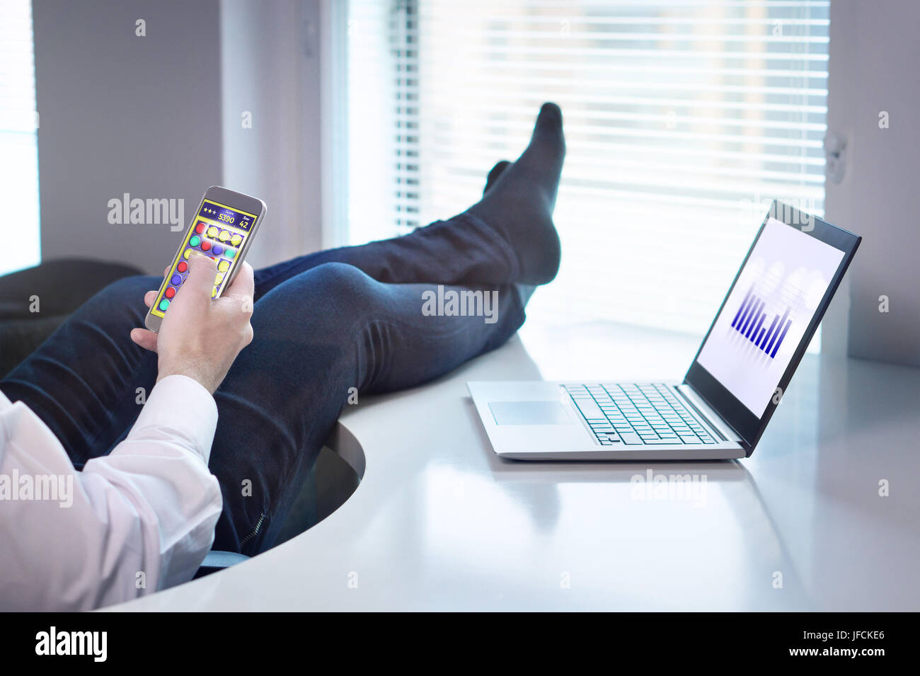 Lazy office worker playing mobile game with smartphone during work hours. Avoiding his job and being lazy with feet and socks on table. Stock Photo