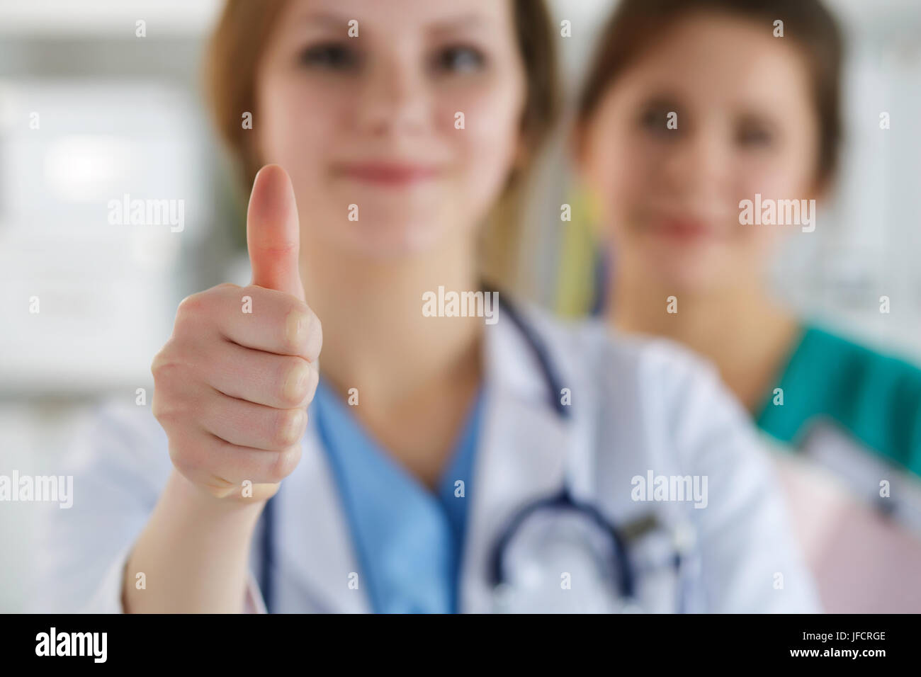 Smiling female doctor showing OK sign with thumb up standing at her office with her colleague on background. High level quality medical service, best  Stock Photo