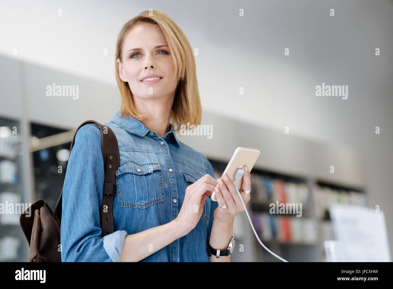 Wait a second. Smiling woman with brooding eyes testing the latest model of a smartphone while shopping for a new technological device at an electroni Stock Photo