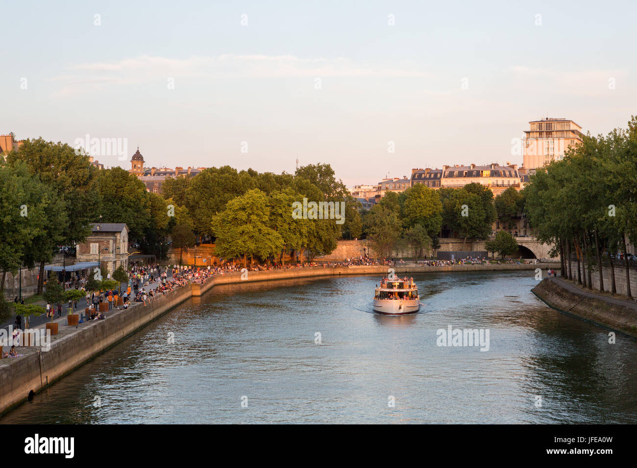 A passenger boat with tourists cruises along the Seine River while Parisians sit along the river banks. Stock Photo