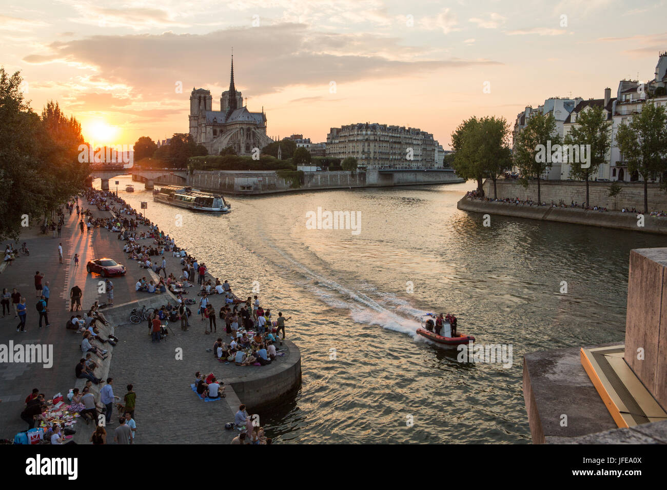 The sun sets near the Notre Dame Cathedral as boats cruise along the Seine and Parisians sit along the river banks. Stock Photo
