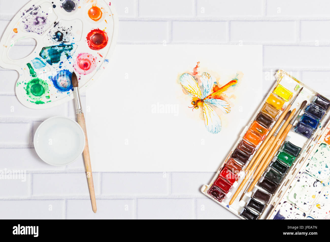 Hand Drawn Sketch of Watercolor Orange Dragonfly,with lying paints, paintbrushes and palette on the white brick background - concept of human creativi Stock Photo